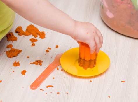 a small children's hand makes an orange sand cupcake on a beige table, a yellow plastic toy plate, playing with sand at home