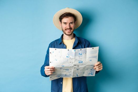 Happy and positive guy on vacation, looking at camera and holding map, smiling excited, going on summer travel, standing on blue background.