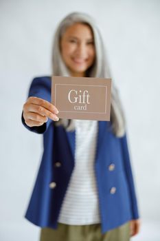 Positive middle aged Asian woman with long silver hair in blue jacket on light grey background in studio, focus on hand with gift card