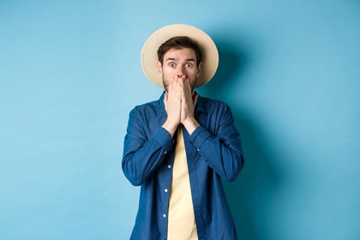 Shocked and alarmed tourist gasping, covering mouth with hands and looking startled at camera, standing on blue background.