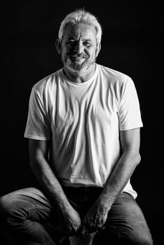 Mature man smiling looking at camera. Senior male with white hair and beard laughing wearing casual clothes isolated on black background. Studio shot in black and white.