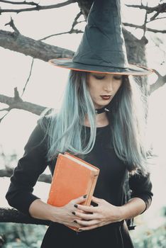 Beautiful young witch holding book of magic in autumn forest. Halloween theme