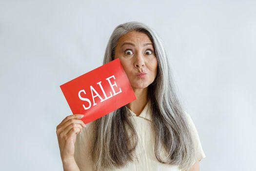 Funny middle aged Asian woman with loose silver hair shows red Sale sign grimacing on light background in studio