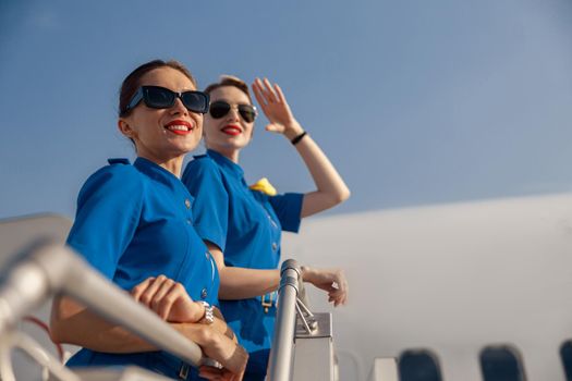 Portrait of two cheerful air stewardesses in blue uniform and sunglasses smiling away, standing together on airstair on a sunny day. Aircrew, occupation concept