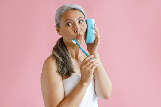 Funny middle aged Asian lady with bath towel holds toothbrush using sponge as phone on pink background in studio. Mature beauty lifestyle