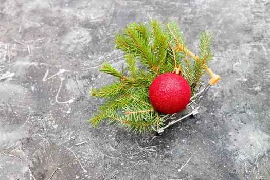 spruce green branch and red shiny Christmas-tree toy ball in a miniature shopping trolley on a black background, copy space