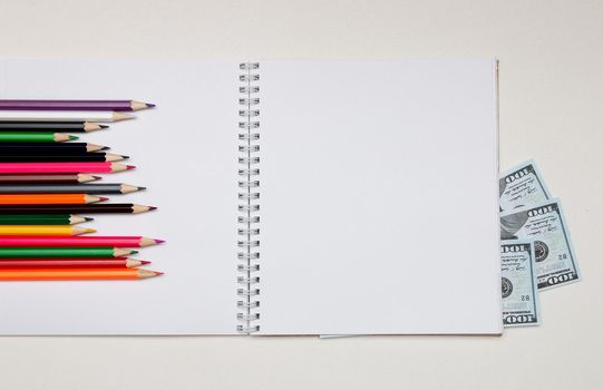 colored pencils lie on a white notebook with a spring and readable sheets of dollar banknotes money on a white table pencil sharpening sharpen color top view spring copybook copy space