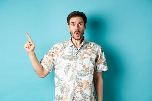 Surprised and amazed tourist in hawaiian shirt pointing finger left, showing promo and staring at camera, standing on blue background.