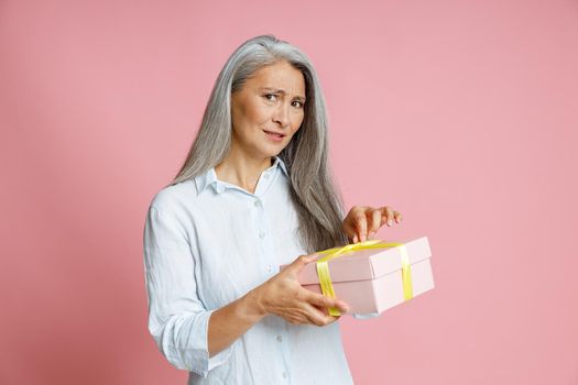 Doubting grey haired Asian woman opens gift box decorated with silk ribbon posing on pink background in studio