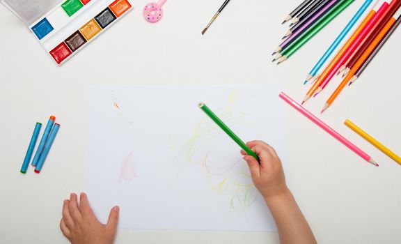 a child draws with crayons on a white lsita on a white table top view place copy child hand holds a green pencil of paint watercolor pen brush wax knives lie next