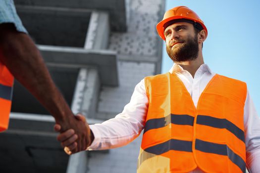 Two men engineers in workwear shaking hands against construction site, close up