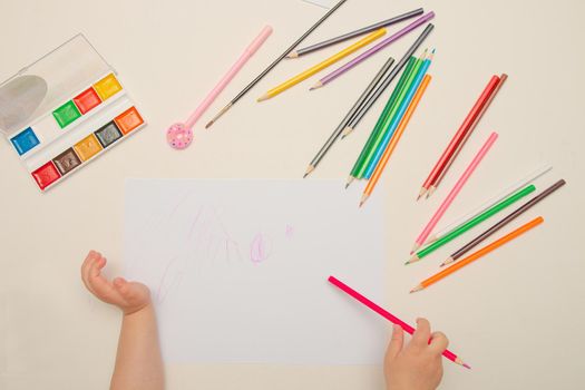 a child draws with crayons on a white lsita on a white table top view place copy child hand holds a Pink pencil of paint watercolor pen brush wax knives lie next