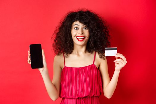 Excited lady in red dress showing plastic credit card and smartphone screen, smiling amazed, standing on studio background.