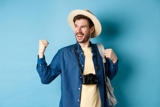 Cheerful tourist rejoicing on vacation, celebrating summer holidays, showing fist pump gesture and saying yes with satisfied face, looking aside, blue background.