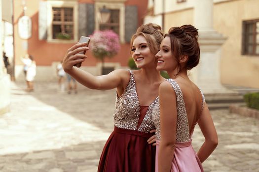 Two attractive european girl friends take selfie on mobile phone outdoors in city. Smiling young women with beautiful haircuts wearing dresses. Sunny daytime. Concept of happy lifestyle