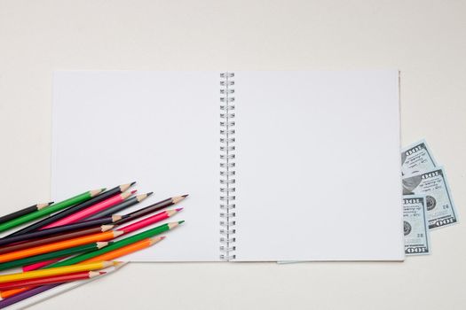 colored pencils lie on a white notebook with a spring and readable sheets of dollars banknotes money on a white table pencil sharpening sharpen color top view spring copybook copy space