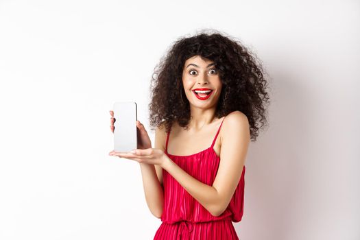 Fashionable lady in red dress and makeup, showing mobile phone screen and smiling, introduce smartphone application, standing over white background.