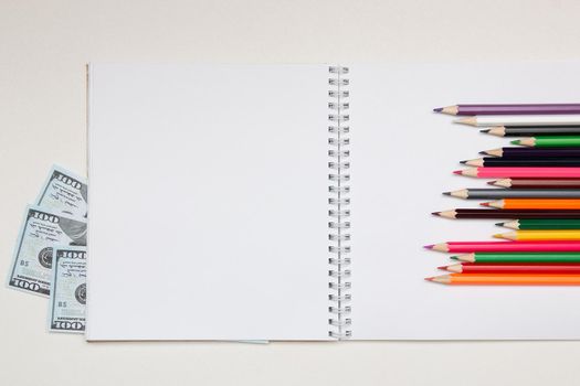 colored pencils lie on a white notebook with a spring and readable sheets of dollars banknotes money on a white table pencil sharpening sharpen color top view spring copybook copy space