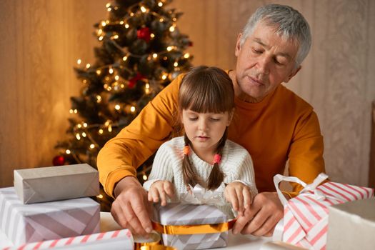Grandfather and granddaughter packing gifts while sitting at table in room with xmas tree on background, child and senior man wearing casual clothing, ties ribbon on bow.