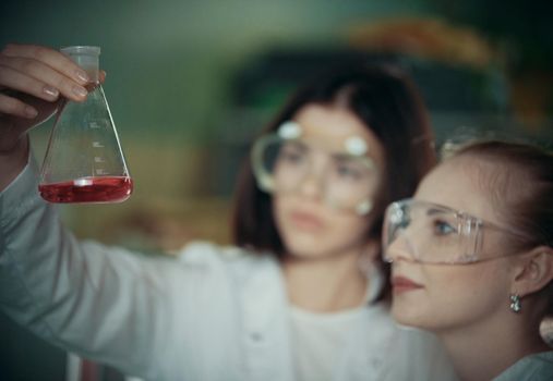 Chemical laboratory. Two young woman holding a flask with red liquid in it. Close up
