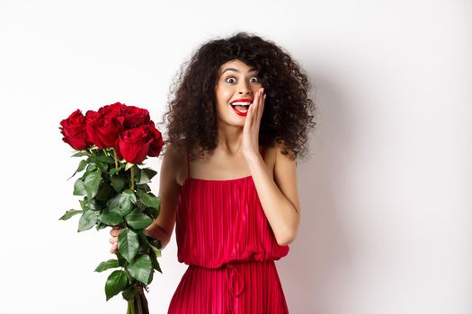 Excited curly woman in red dress, receive bouquet of roses and look surprise, rejoicing from romantic gift, standing on white background.