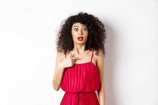 Surprised young woman in red dress pointing at herself, being confused and shocked, standing on white background. Copy space
