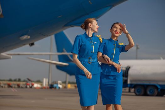 Two cheerful air stewardesses in bright blue uniform walking outdoors in front of passenger aircraft on a sunny day. Occupation concept