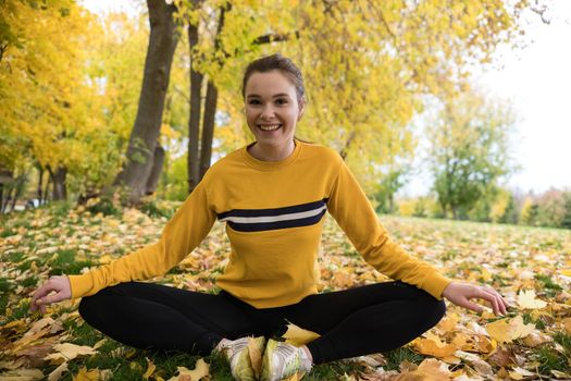 Autumn photo of sporty woman stretching in forest at morning against background of trees. Looking to the camera. Telephoto shot