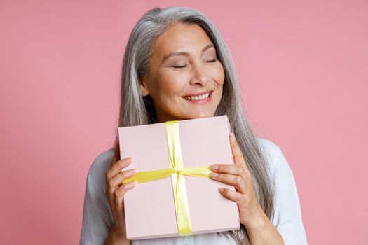 Cheerful mature Asian lady with long loose hair holds gift box decorated with ribbon posing on pink background in studio
