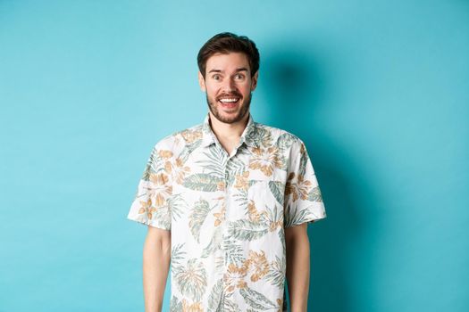 Summer holiday. Handsome happy man in hawaiian shirt looking amused, smiling at camera, standing on blue background.