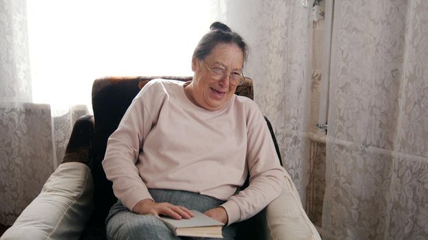 An elderly woman sitting in a chair, holding a book, laughing. Portrait