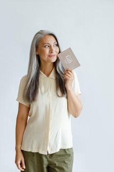 Thoughtful hoary haired Asian lady customer holds gift card standing on light grey background in studio. Shopping certificate