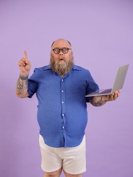 Funny mature man with overweight in tight blue shirt got interesting idea holding modern laptop on purple background in studio