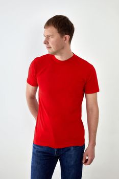 Glad young male in jeans and red t shirt standing with hand behind back against white background and looking away