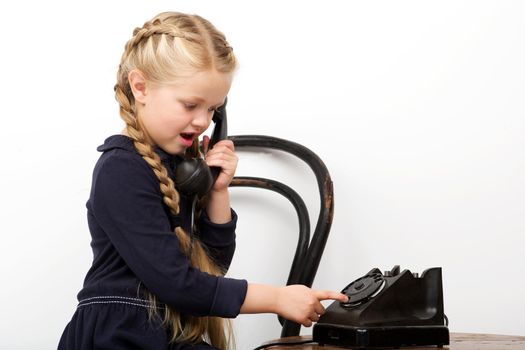 Girl sitting on chair and talking by old phone. Charming stylish kid posing in vintage room interior with old furniture. Joyful girl with braids wearing stylish fashionable clothes