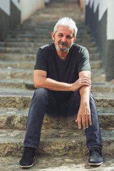 Mature man sitting on steps in the street. Senior male with white hair and beard wearing casual clothes in urban background.