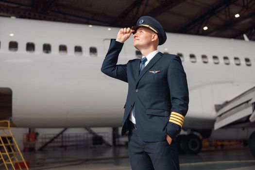 Inspired pilot in uniform looking away, adjusting his hat, standing in front of big passenger airplane in airport hangar. Aircraft, occupation, transportation concept