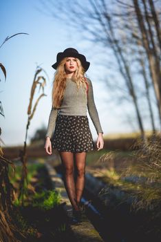 Beautiful young blonde woman, model of fashion, in rural background. Girl wearing sweater, skirt and hat.