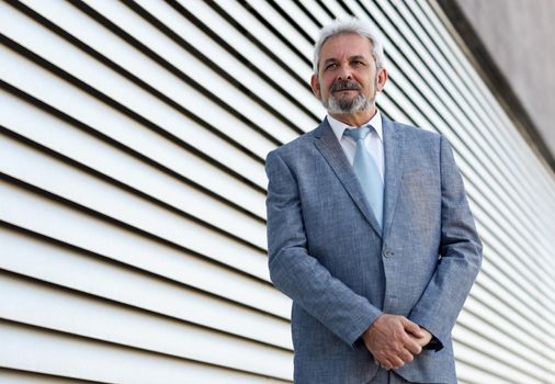 Portrait of a senior businessman with arms crossed outside of modern office building. Successful business man wearing formal suit and tie smiling in urban background.