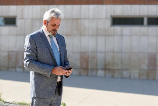 Portrait of a senior businessman texting with a smart phone outside of modern office building. Successful business man in urban background.