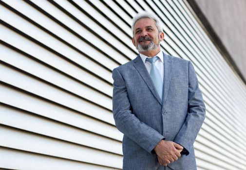 Portrait of a senior businessman with arms crossed outside of modern office building. Successful business man wearing formal suit and tie smiling in urban background.