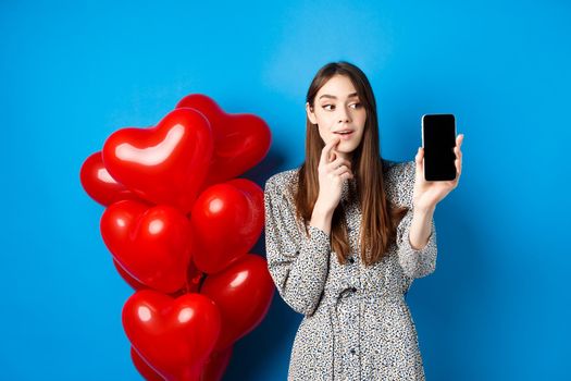 Valentines concept. Beautiful girl looking and showing mobile screen, checking out online shopping offer on lovers day, standing on blue background.
