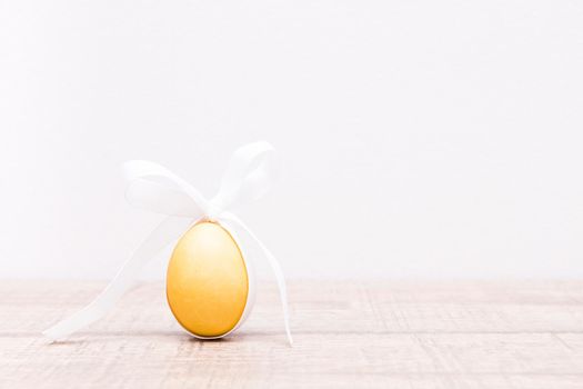 yellow easter egg with a white ribbon tied in a bow in the form of rabbit ears on a light phone, top view, copy space, texture background, wooden table