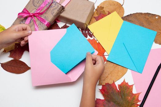 children's hand puts a card in an envelope, bright colors, gifts with beautiful ribbons, greetings for the holiday, Thanksgiving, birthday, autumn leaves on a white table, copy space top view, pink, yellow, blue