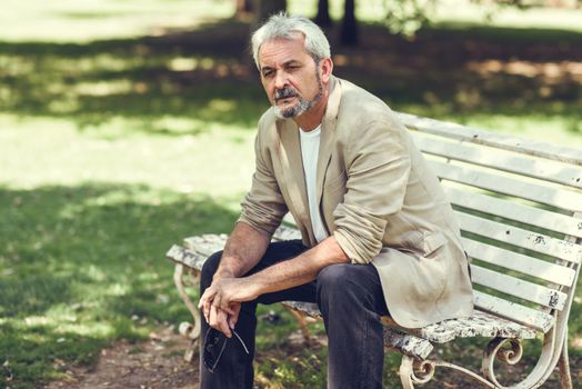 Portrait of a pensive mature man sitting on a bench in an urban park. Senior male with white hair and beard wearing casual clothes.