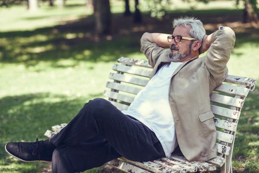 Portrait of a pensive mature man sitting on a bench in an urban park. Senior male with white hair and beard wearing casual clothes and eyesglasses.