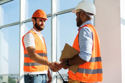 Two men engineers in workwear shaking hands against construction site, close up