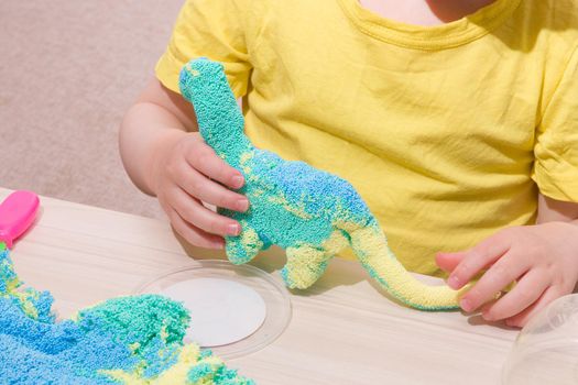 the child plays plasticine from polymer granules at the table, the original plasticine from tiny balls, the child plays with a dinosaur from plasticine, what to play with the child at home