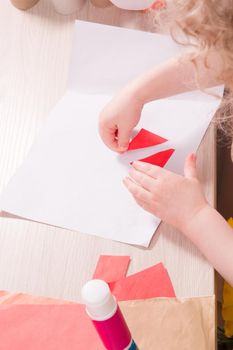 a small child glues paper, children's safe scissors and colored paper on the table, what to do with the child at home, children's hands glue colored triangles on a sheet of white paper