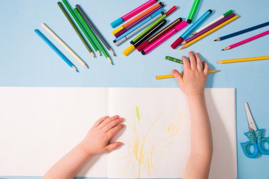 a small child draws with pencils in an album, copy space, top view, blue background, pencils, wax crayons, scissors and the child's hands on the table